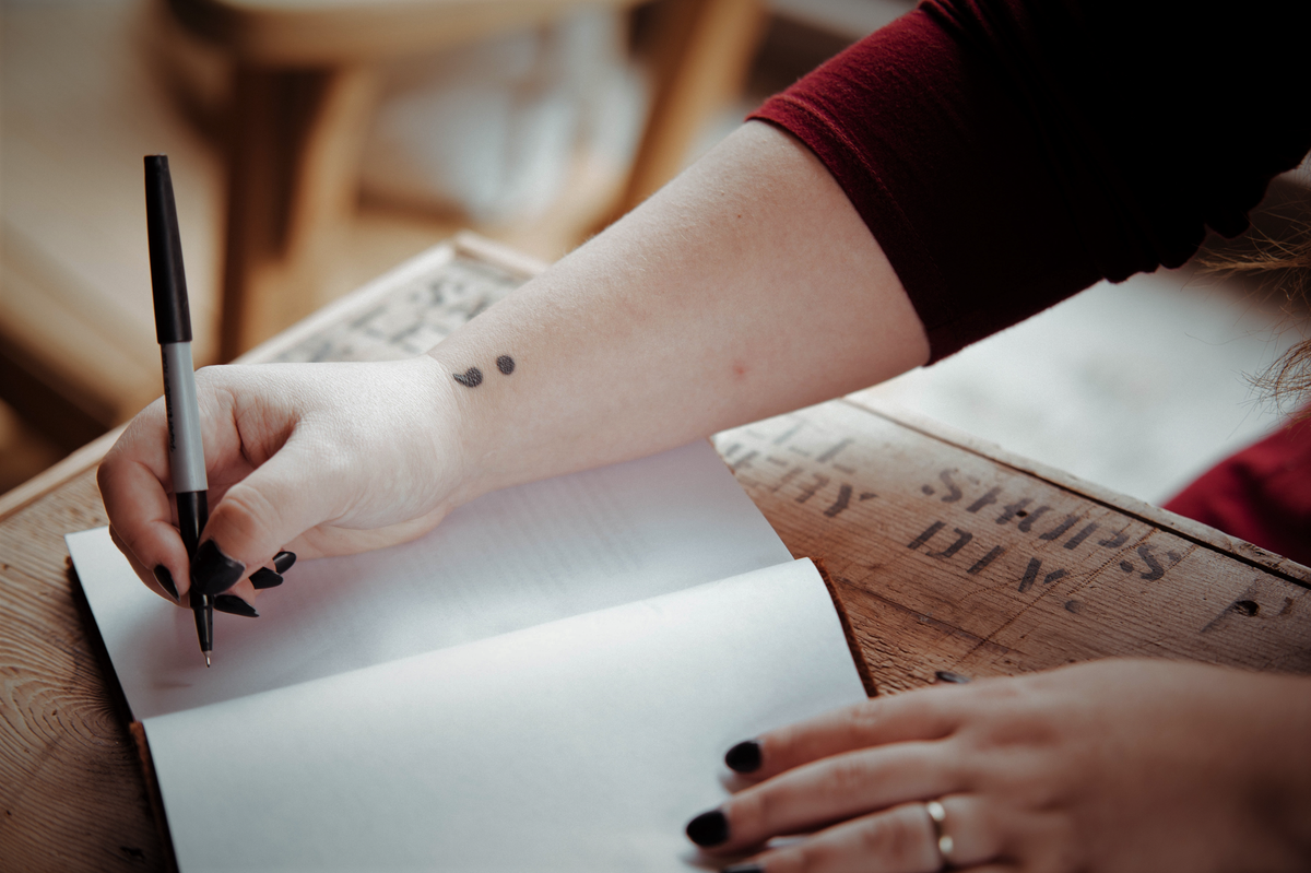 Woman writing in a blank notebook with a semicolon tattoo on her wrist