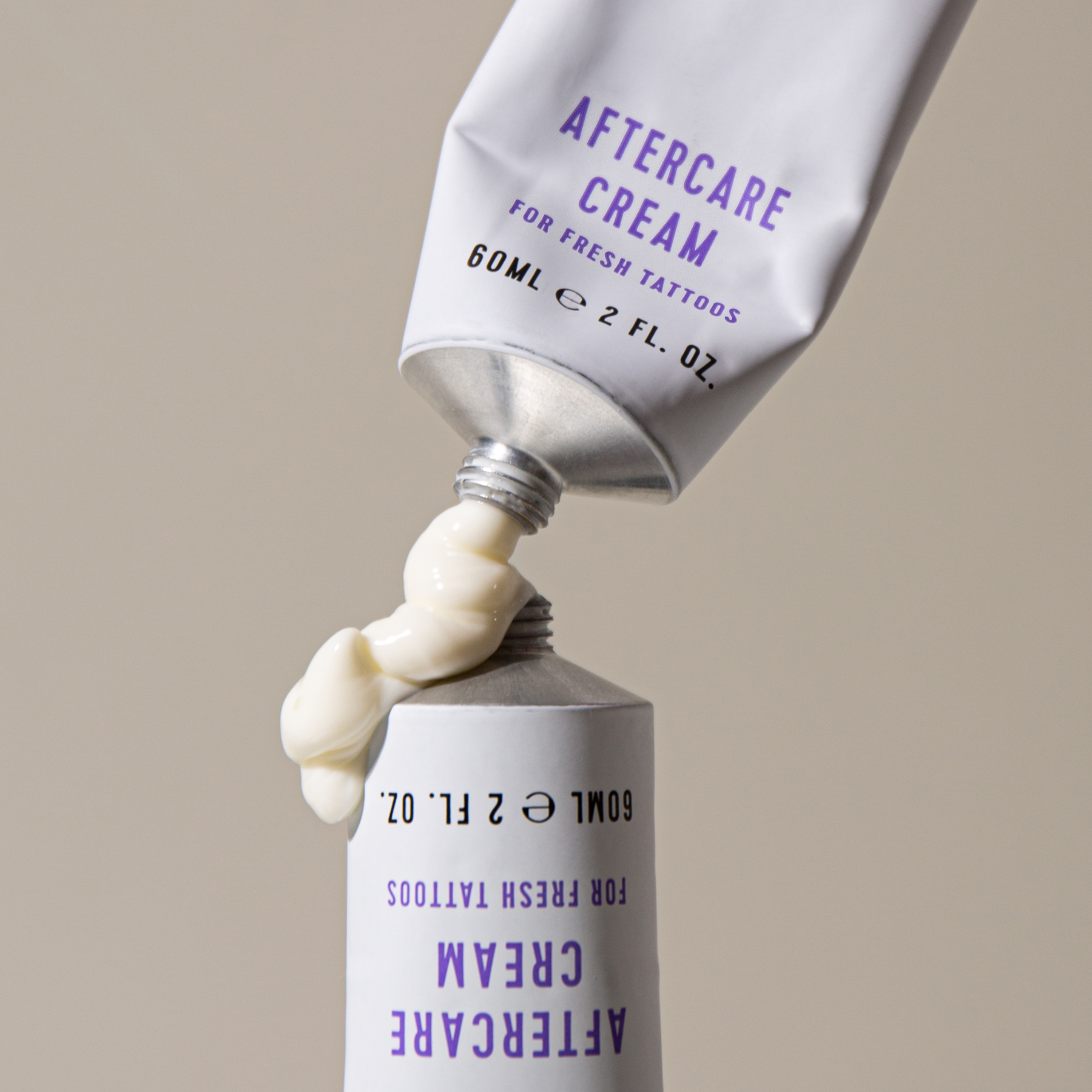 A white tube labeled "AFTERCARE CREAM FOR FRESH TATTOOS" is being squeezed, dispensing a swirl of white cream. The text on the tube also includes “60ML e 2 FL. OZ.” in black and purple. The cream is emerging smoothly from the open tube, with the bottom of the tube visible. The context is minimal, featuring a plain, neutral background that draws focus to the action of the cream being squeezed out.